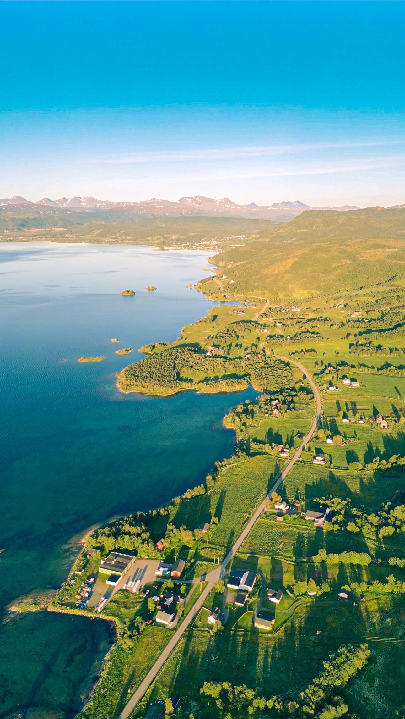 En luftfoto av en vakker kystlinje med grønne områder, hus og fjell i bakgrunnen.