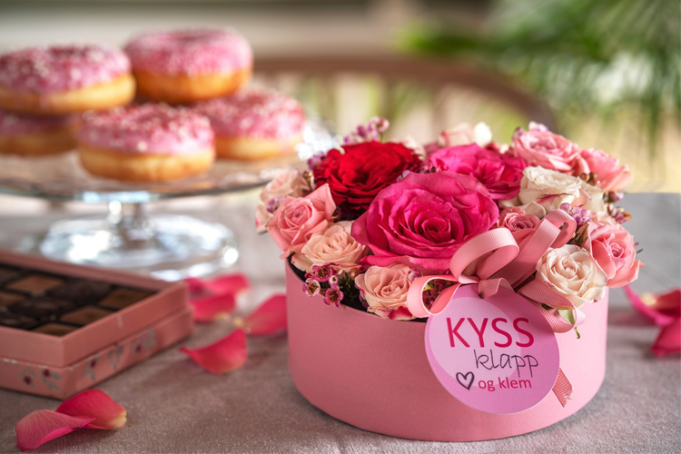En rosa blomsterboks med røde og rosa roser, med donuts i bakgrunnen og en sjokoladeeske ved siden av.