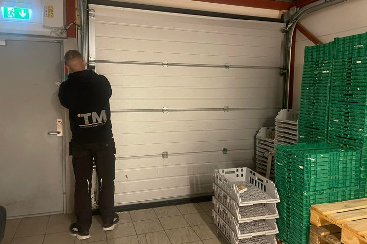 En person lukker en garasjeport i et lager med grønne plastkasser stablet ved siden av.