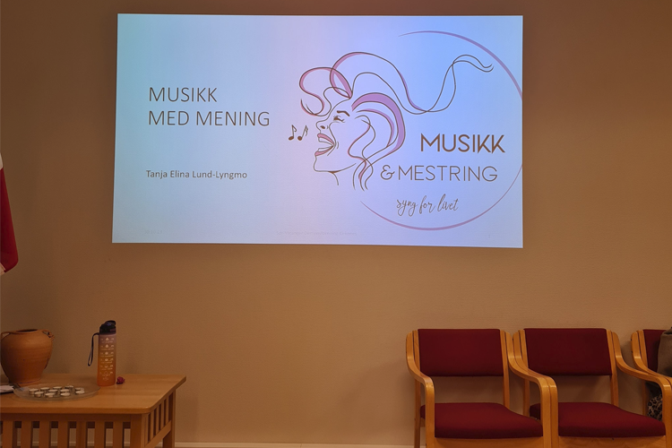 Presentasjon om musikk med mening, med logo for Musikk & Mestring på veggen. Stoler i rommet.