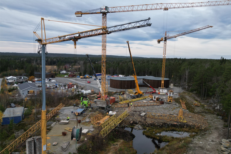 Byggeplass med flere kraner, lastebiler og maskiner i et skogsområde, med bygninger i bakgrunnen.