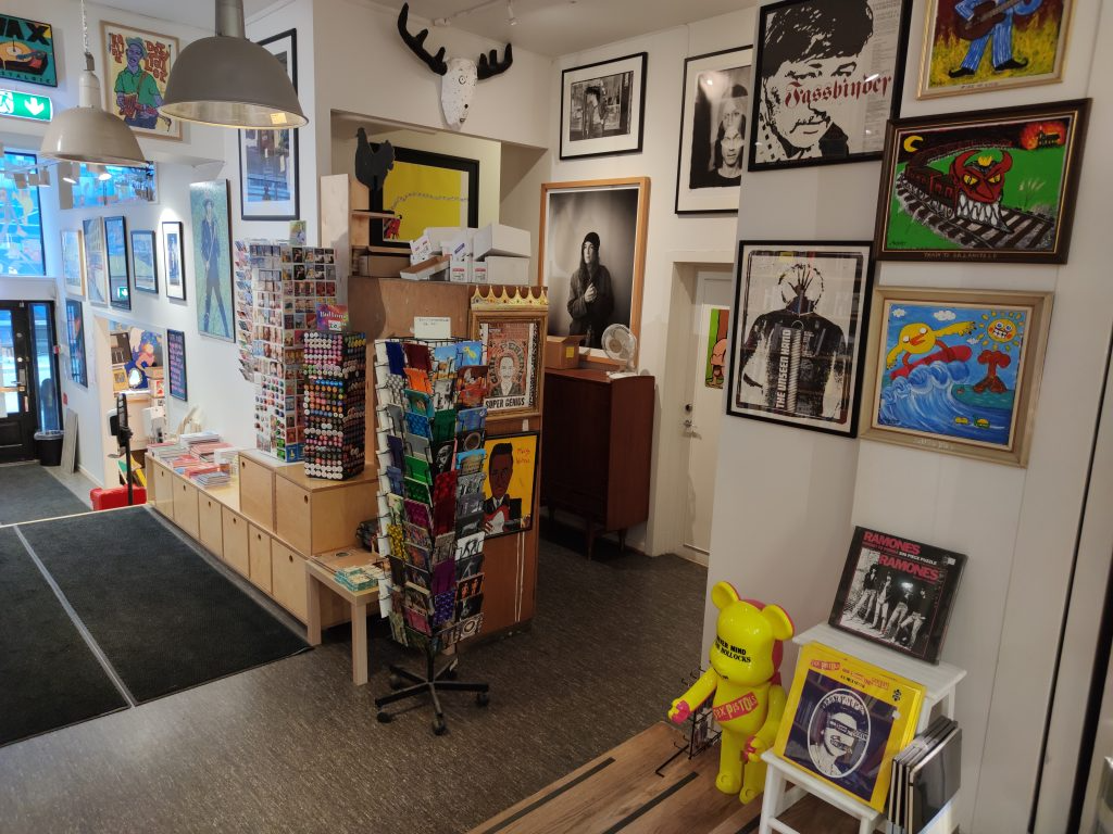 En kunstgalleriinnredning med fargerike malerier, plakater, og en utstillingsvegg med kunstverk og samlerobjekter.