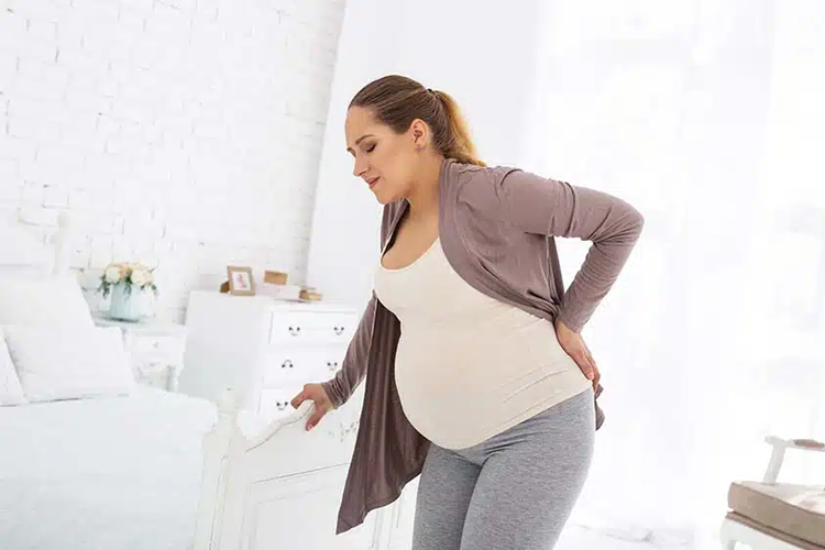 Gravid kvinne står ved en seng og holder seg til møbelet, ser ut til å oppleve ubehag i ryggen.