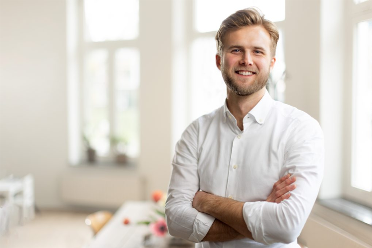 En smilende mann med skjegg står med armene krysset i et lyst rom med vinduer.