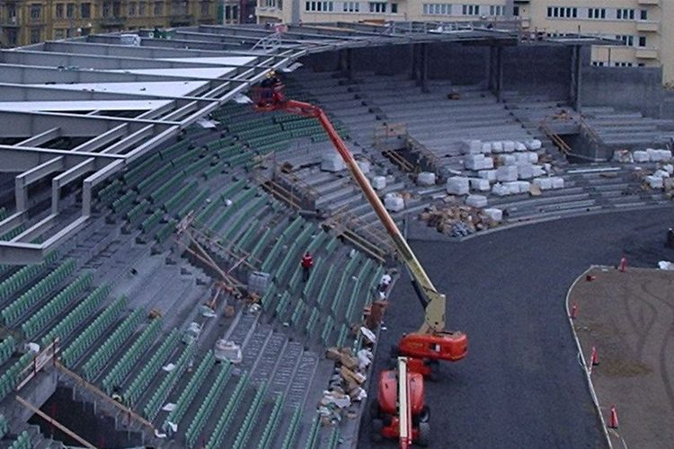 Byggearbeid på en stadion med stillaser, heiser og grønne seter i bakgrunnen.