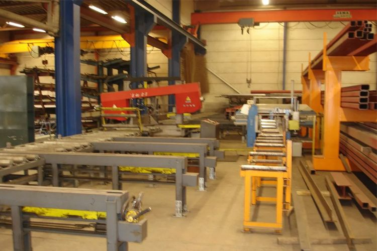 Verksted med maskiner og stålprofiler, inkludert en rød kran og arbeidsbenker i metall.