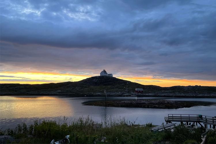 Solnedgang over en øy med et hvitt fyrtårn og en brygge i forgrunnen. Himmel med skyer i varme farger.