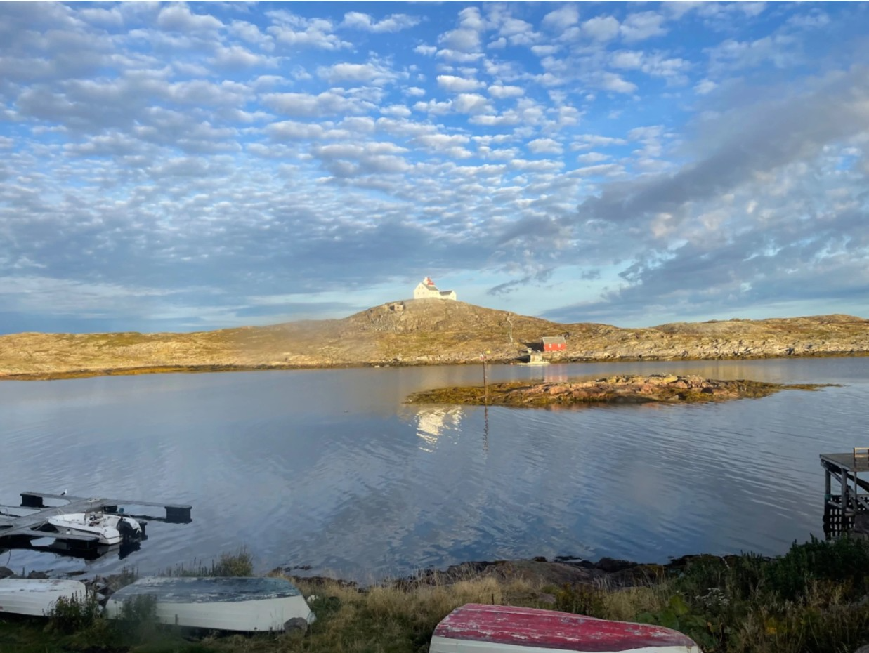 En rolig havn med båter, en øy i midten, og en fyrtårn på toppen av en høyde under en skyet himmel.
