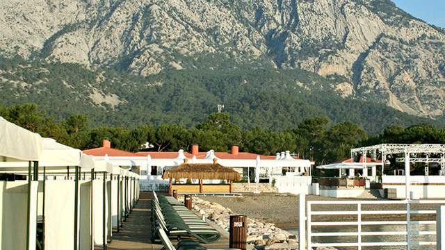 Strandpromenade med solsenger og hvite telt, med fjell i bakgrunnen og bygninger i kystlandskapet.