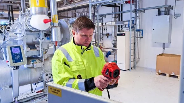 En mann i gul sikkerhetsjakke bruker en måleinstrument i et industrielt miljø med rør og utstyr.