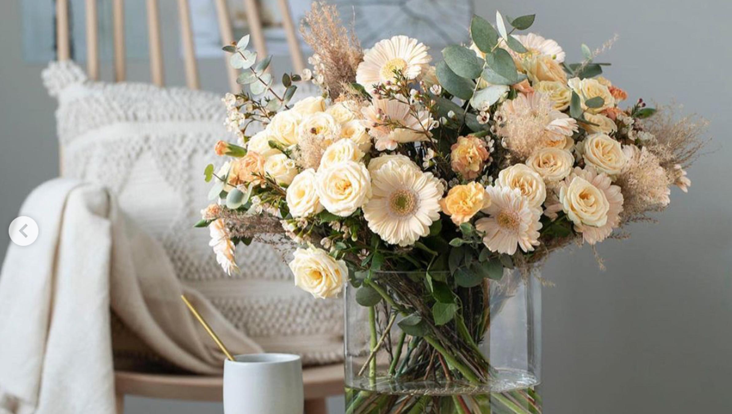 En vakker bukett med kremfargede roser og tørkede blomster i en glassvase, plassert ved en stol.