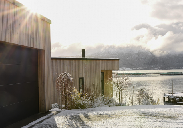Moderne trehus ved vannet med snø og fjell i bakgrunnen, solstråler som skinner over landskapet.