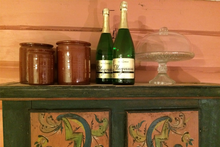 To flasker musserende vin og to brune krukker på et dekorert skap med en glasskakefat.
