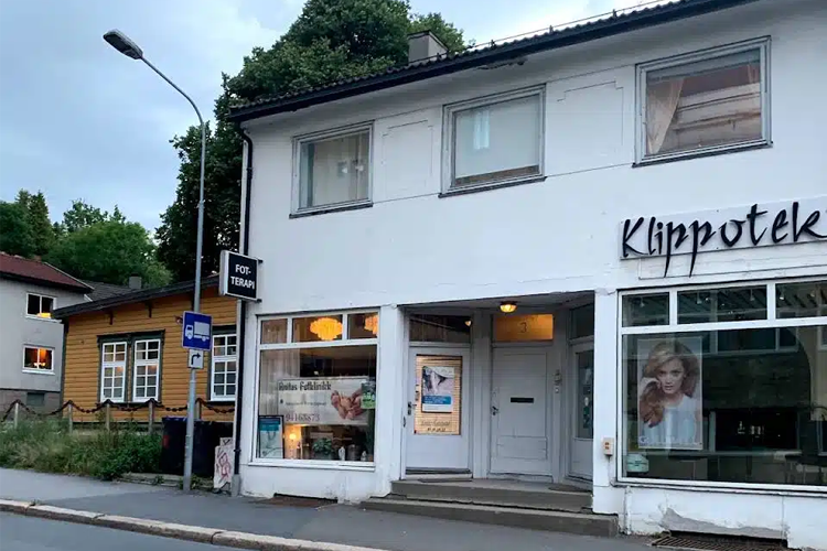 En frisørsalong med store vinduer og en inngangsdør, beliggende i en bygate med trær i bakgrunnen.