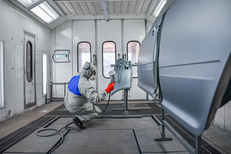 En person i beskyttelsesdrakt sprayer bilkarosseri i et malerom med hvite vegger.