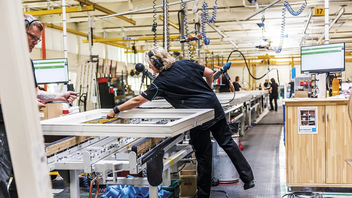 En fabrikk med arbeidere som monterer vindusrammer på en produksjonslinje.