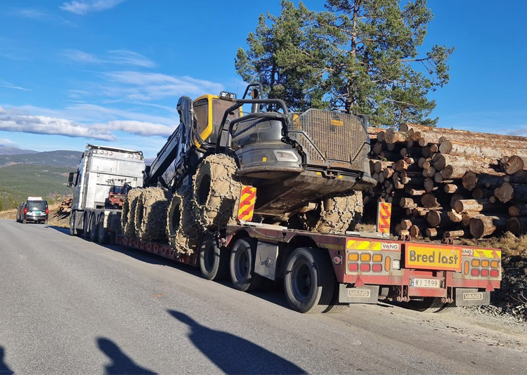 En lastebil med en stor gravemaskin lastet med gjørme, parkert langs en vei med tømmerstokker i bakgrunnen.