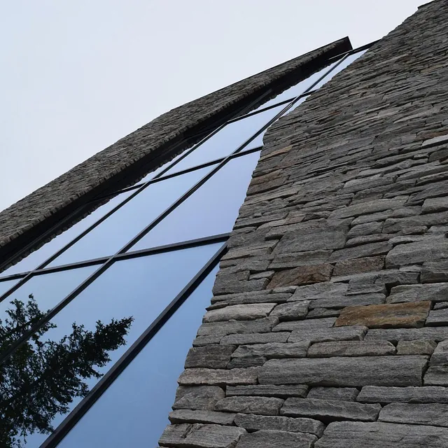 Moderne bygning med steinvegg og store vinduer som reflekterer trær i nærheten.