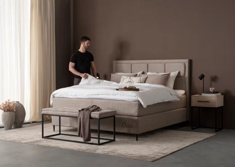 En mann retter opp sengen i et moderne soverom med beige farger og stilige møbler.