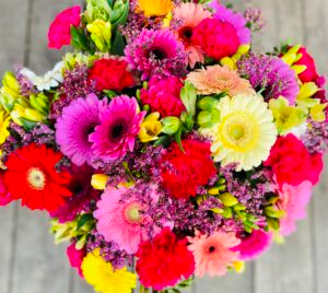 En fargerik bukett med blomster, inkludert gerbera, nelliker og liljer, på en treoverflate.