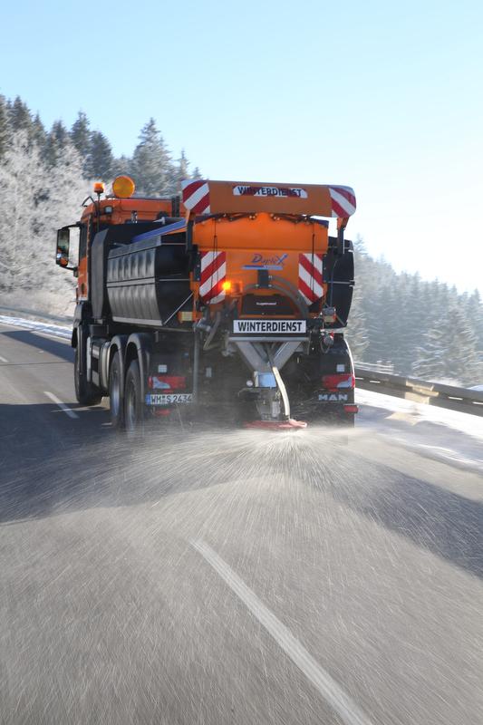 En snøplogbil sprer salt på en vintervei med snødekte trær i bakgrunnen.