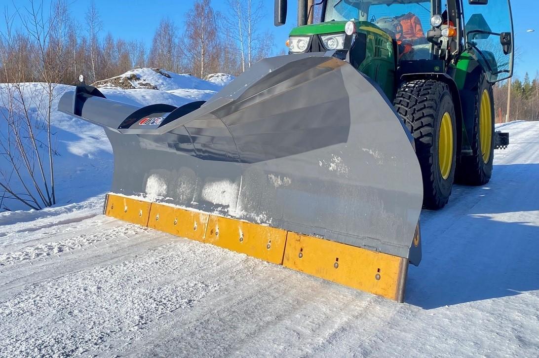En snøplog festet til en traktor som rydder snø fra en vei i solfylt vintervær.