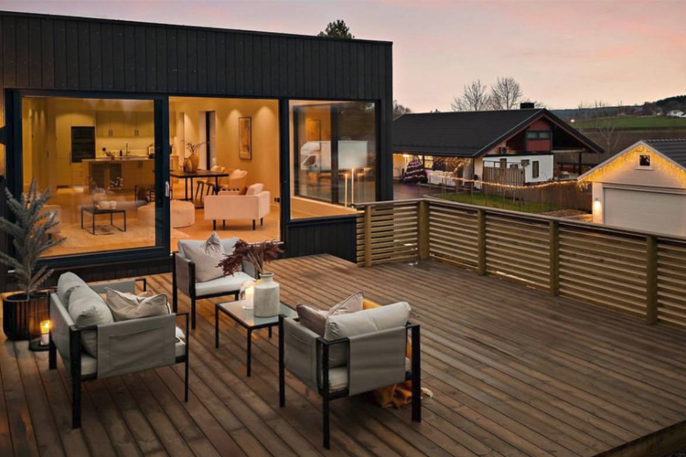 Moderne terrasse med stoler og bord, med utsikt til et stilrent hus og solnedgang i bakgrunnen.