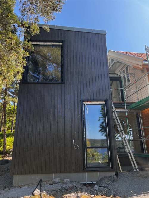 Moderne, svart trehus med store vinduer, plassert i et naturskjønt område med trær og en byggeplass i bakgrunnen.
