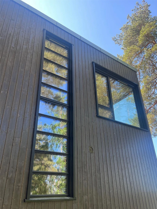 Moderne husvegg med store vinduer og trepanel, omgitt av grønne trær under en klar blå himmel.