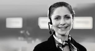Kvinne med headset smiler i en kontormiljø, med uskarpe bakgrunner og moderne teknologi.