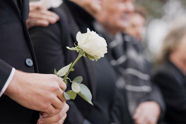 En person holder en hvit rose, mens flere mennesker står sammen i bakgrunnen under en minneseremoni.