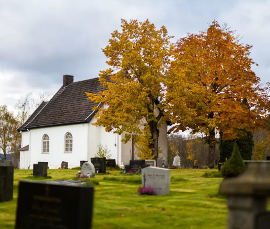 Hvit kirke med høstfargede trær i bakgrunnen, omgitt av gravsteiner på en rolig kirkegård.