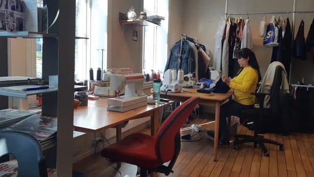 En kvinne sitter ved et bord i et rom med symaskiner, klær og hyller med materialer.