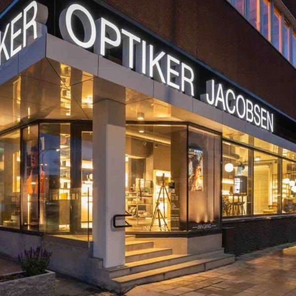 Optiker Jacobsen butikkfasade med store vinduer og lys, beliggende i en moderne bygning.