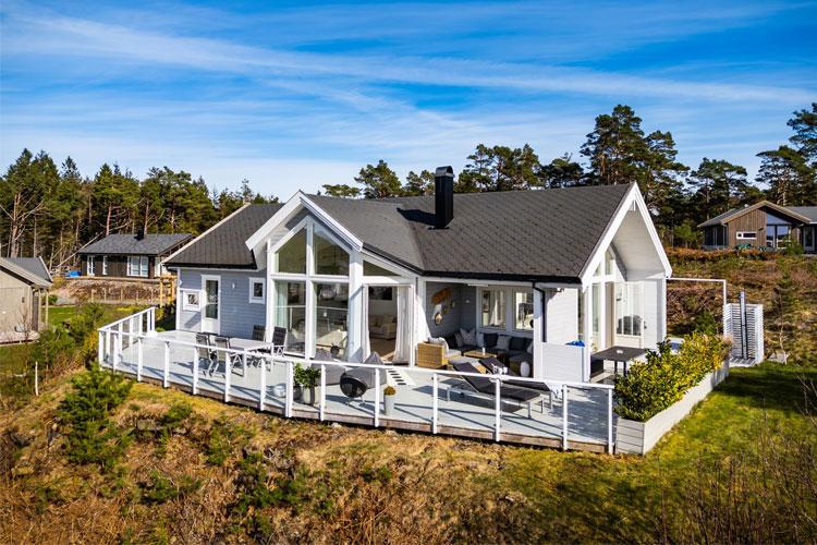 Moderne enebolig med stor terrasse, beliggende i naturskjønne omgivelser med trær og blå himmel.