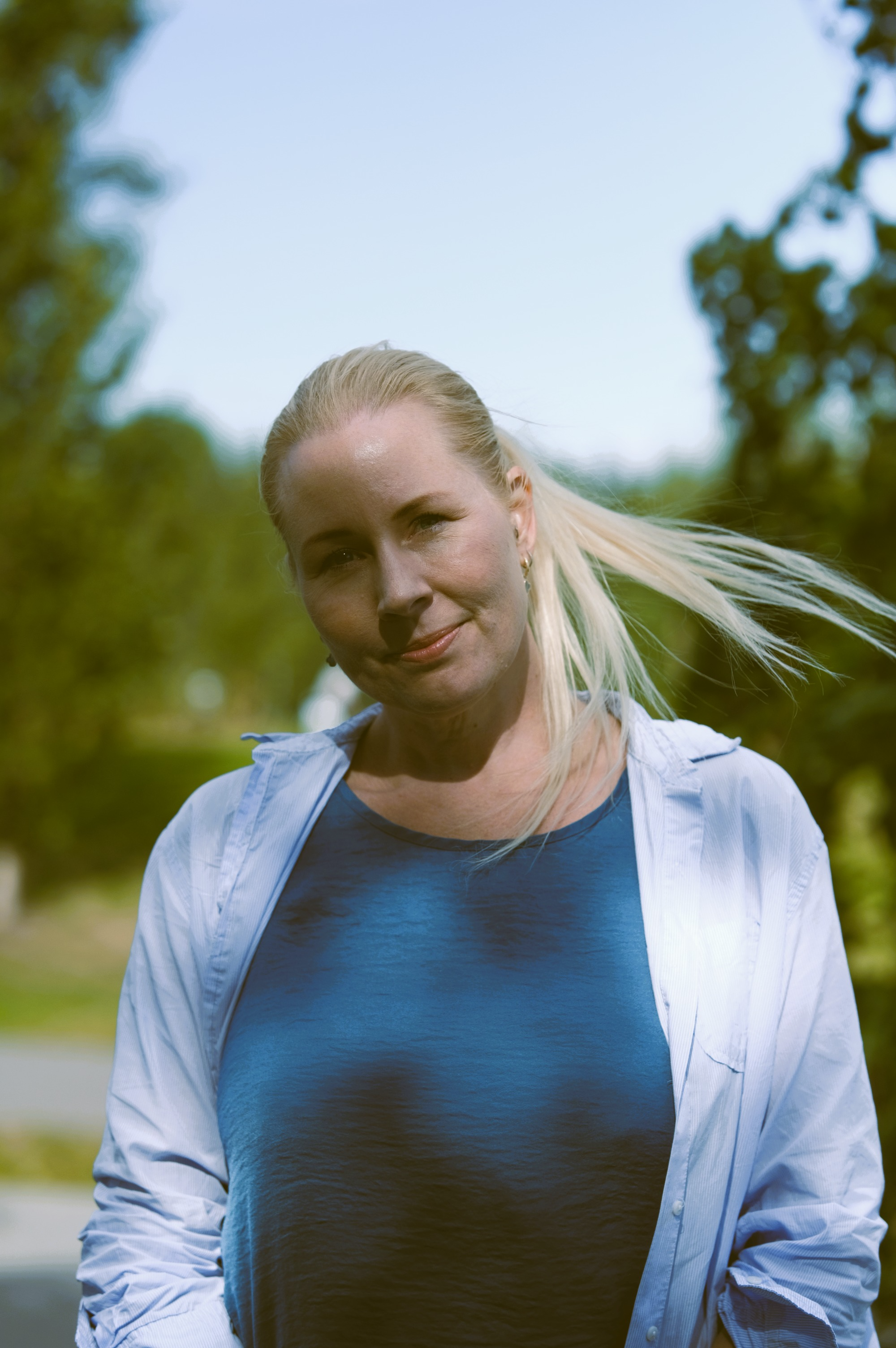 Kvinne med langt, lyst hår iført en blå topp og hvit skjorte, står ute i solfylt vær.