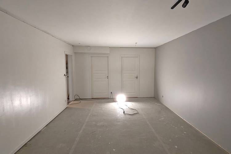 Tomt rom med grå og hvite vegger, to dører og en lampe som lyser opp gulvet.