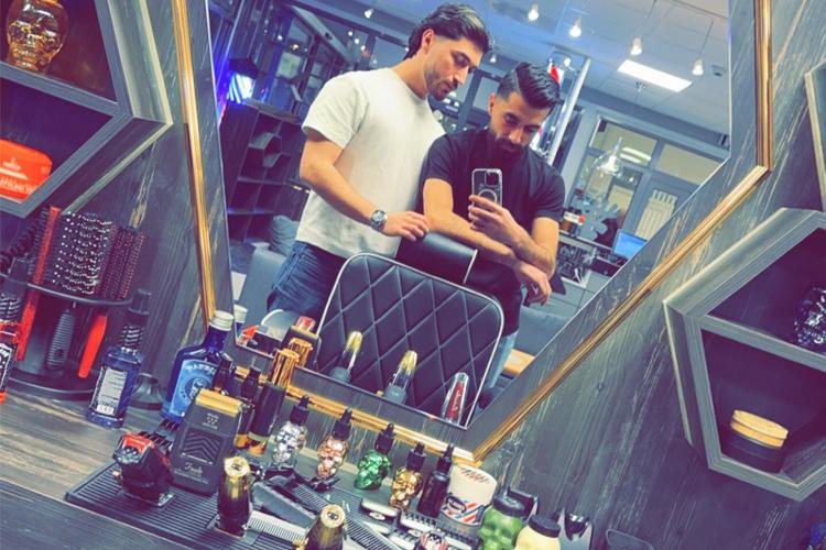 To frisører står foran et speil i en barbersalong, med produkter og verktøy synlige på bordet.