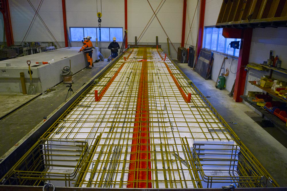 Bilde av en stor betongform med armeringsjern i en fabrikk, med arbeidere som jobber i bakgrunnen.