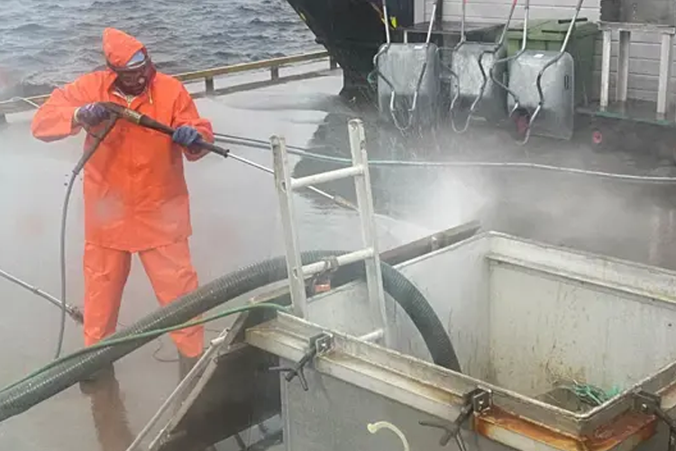 Fisker i oransje regntøy som høytrykkspyler en båt på kaien, med vannsprut og damp.