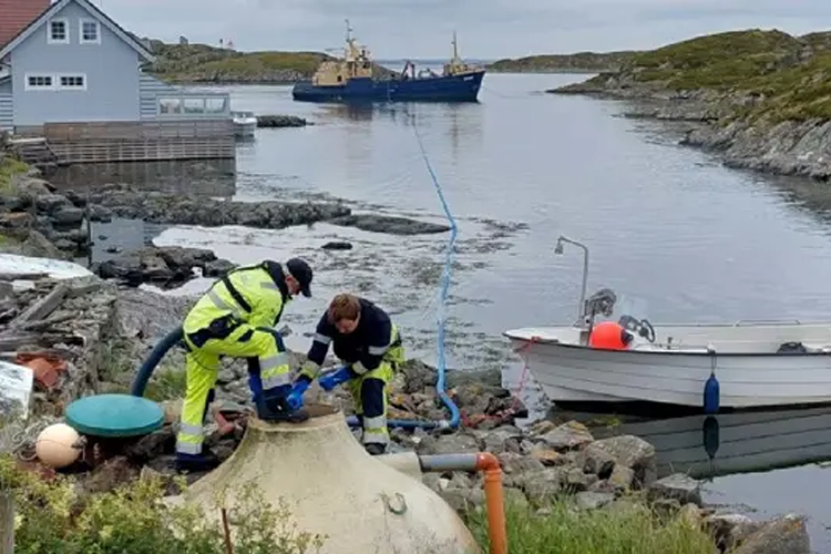 To personer i arbeidsklær jobber med utstyr ved vannkanten, med en båt og en fiskebåt i bakgrunnen.