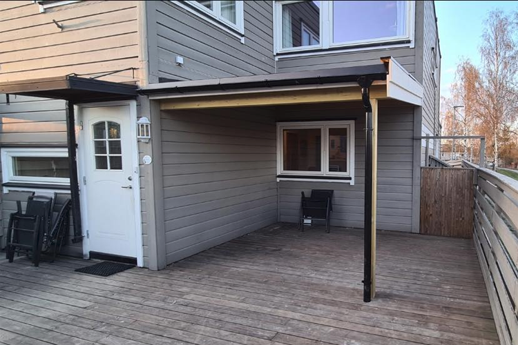 En terrasse med tredekke, en dør og et vindu, plassert ved siden av en grå husvegg.