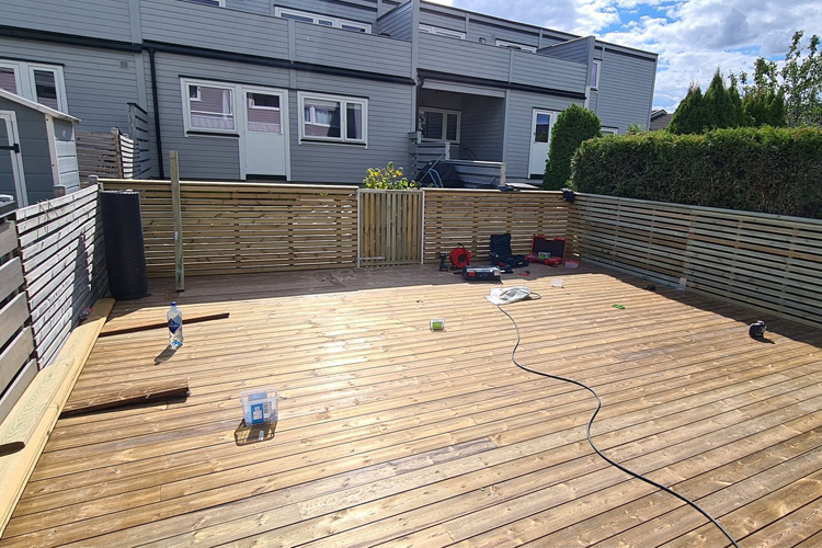 Terrasse med treplanker, gjerde og verktøy, med bygninger i bakgrunnen og sollys som skinner.