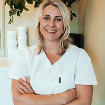 En smilende kvinne i hvit arbeidsdrakt står med armene krysset, omgitt av planter.