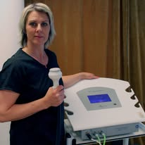 Kvinne med behandlingsapparat i klinikk, holder en håndholdt enhet og ser mot kameraet.