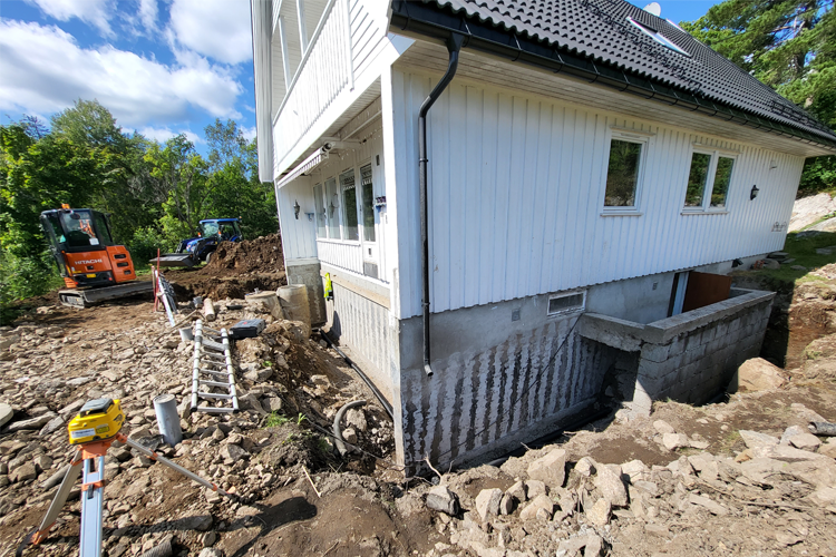 Hus med graving rundt fundamentet, gravemaskin og byggeutstyr i bakgrunnen, og stein og jord på bakken.