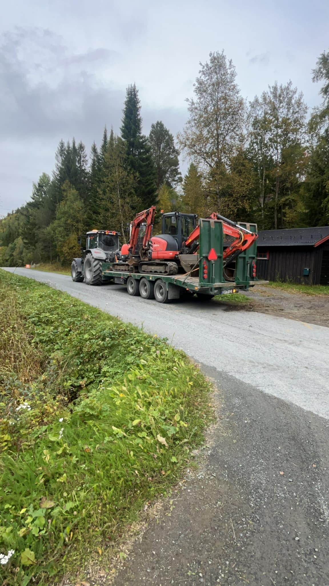 Traktor med gravemaskin på tilhenger kjører langs en grusvei omringet av trær og natur.