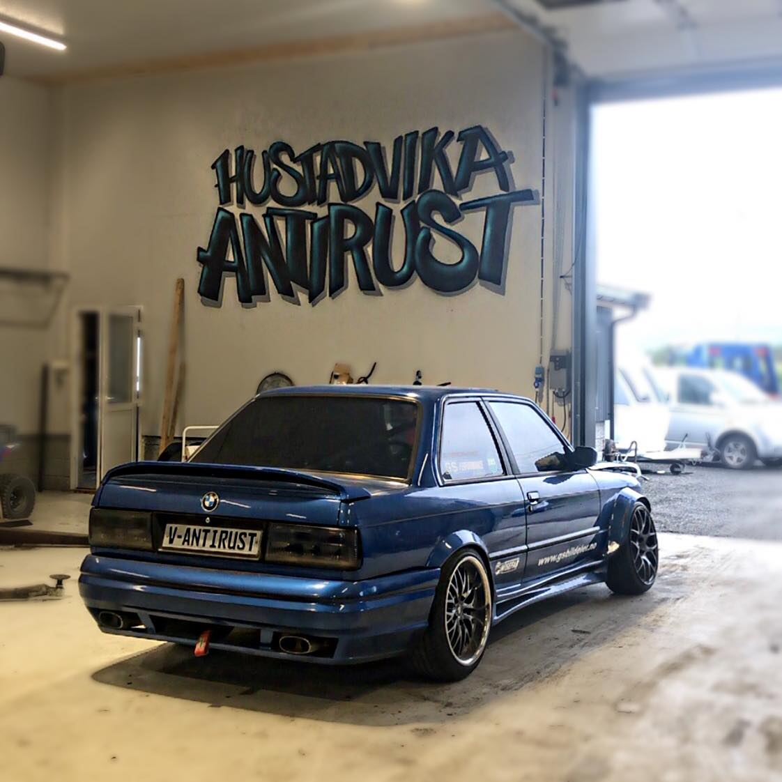 Blå bil med lav profil og svarte felger, parkert i en garasje med graffiti på veggen.