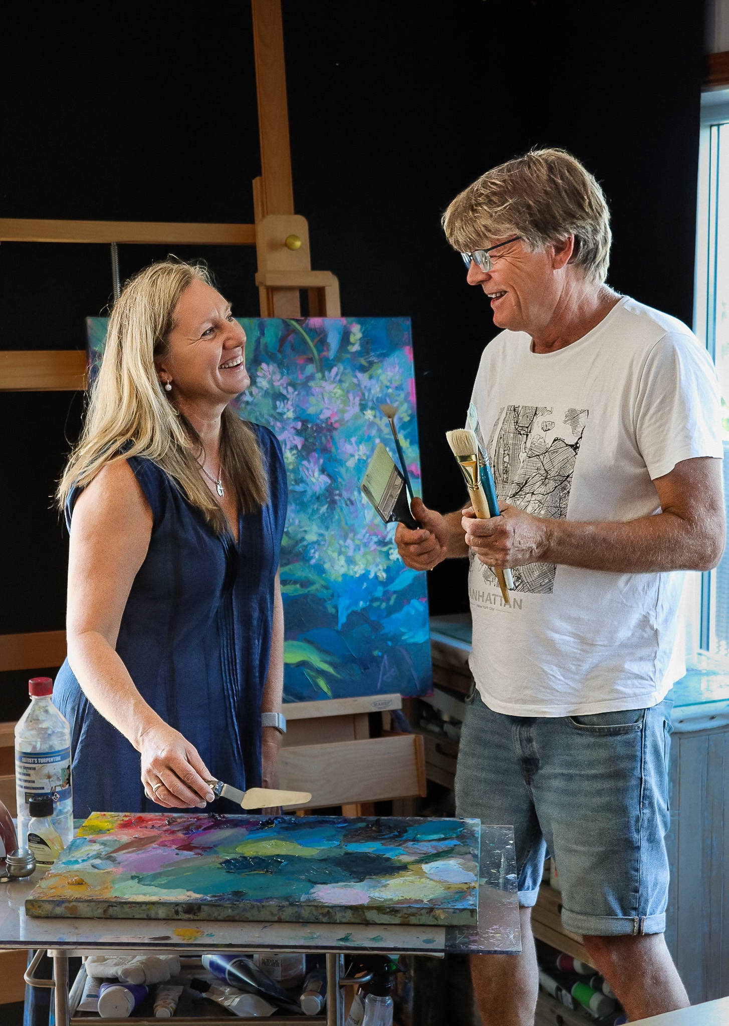 To personer smiler og maler sammen i et kunststudio, omringet av fargerike malerier og kunstutstyr.