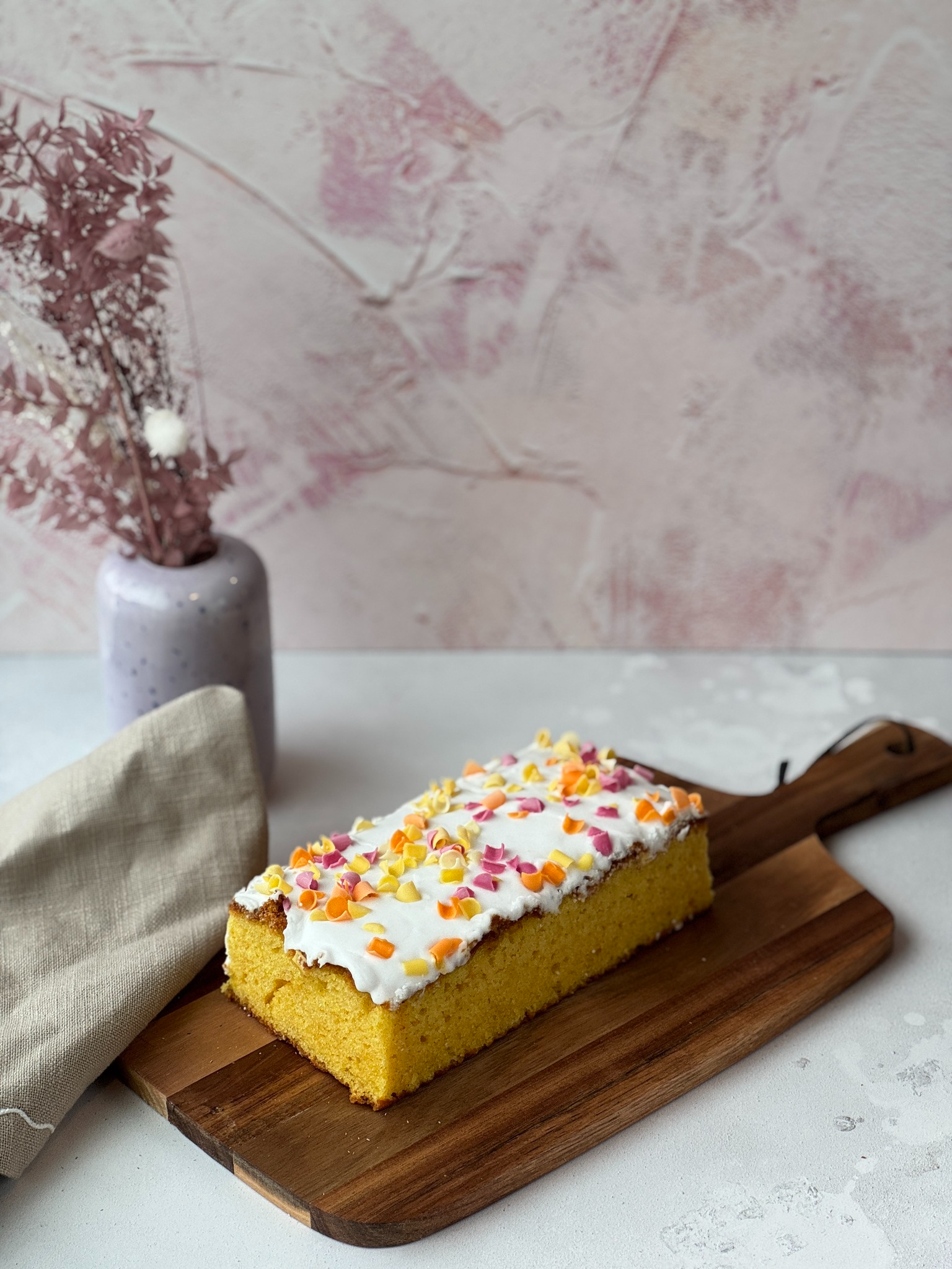 En saftig kake med hvit glasur og fargerike strøssel, servert på en treplate med en vase i bakgrunnen.